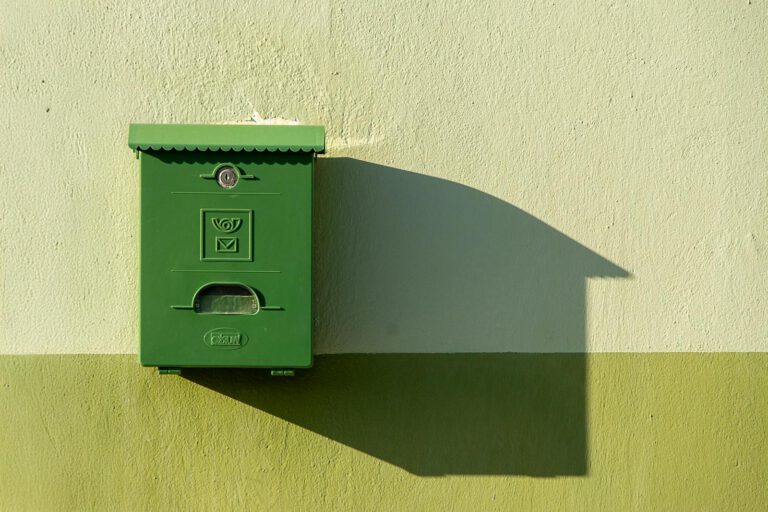 A green postbox mounted on a wall casting a long shadow in bright sunlight.